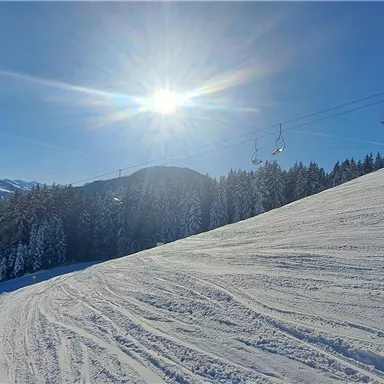 A snow-covered slope under a bright blue sky. The sun is shining brightly and the mountains in the background are visible.