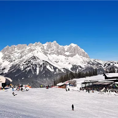 A picturesque winter landscape with snow-covered mountains and a clear blue sky. Skiers and two mountain huts are visible in the foreground.