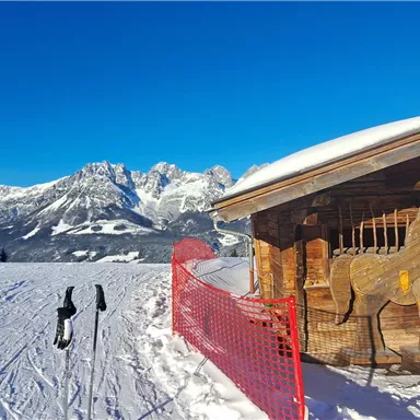 A snow-covered landscape with a wooden building and red barriers. In the background, the mountains and a clear blue sky can be seen.