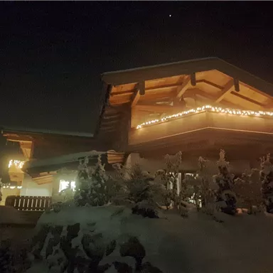 A cozy wooden house in the snow, decorated with lights. It radiates warmth in the cold night.