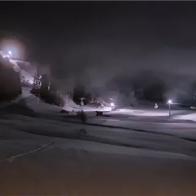 A snowy landscape at night, illuminated by warm lights. In the background, mountains and a ski slope can be seen.