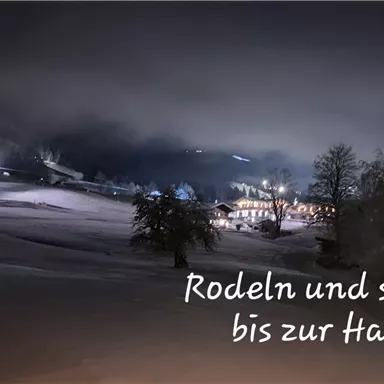 A snowy landscape at night with gentle hills and illuminated paths. The text suggests sledding and skiing right to the doorstep.