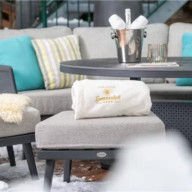 Ein gemütlicher Sitzbereich mit grauen Möbeln und bunten Kissen. Auf dem Tisch steht ein Sektkübel und eine kuschelige Decke.