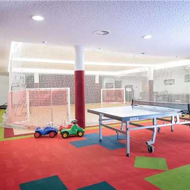 Ein heller Spielraum mit einem Tischtennistisch und bunten Teppichen. Im Raum gibt es auch zwei kleine Spielzeuge und Fußballtore.