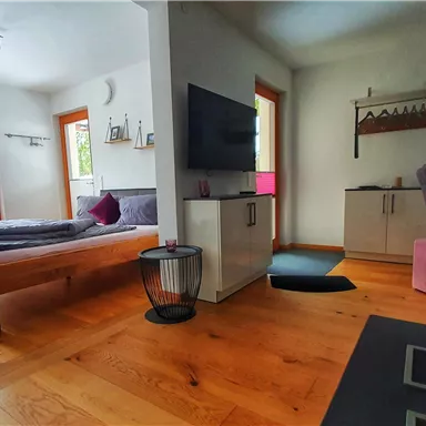 A cozy room with a large window and a TV on the wall. The wooden floor and furniture create an inviting atmosphere.