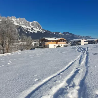 A snowy landscape with mountains in the background and clear blue sky. In the foreground, the tracks in the snow and some buildings can be seen.