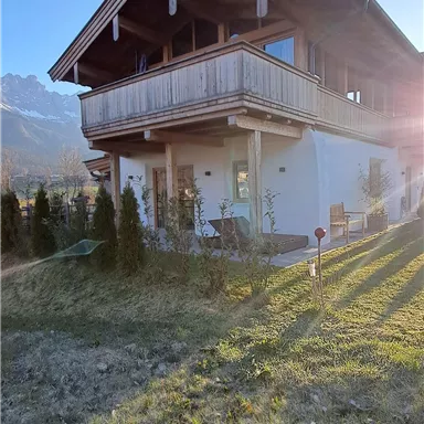 A modern house with a balcony and surrounding garden. In the background, mountains and a clear sky can be seen.