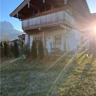 A charming wooden house with a balcony near the mountains. The sun is shining and brightening the garden.