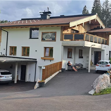 Ein modernes Haus mit mehreren Etagen und einem großen Balkon. Im Vordergrund sind parkende Autos und eine grüne, gepflegte Umgebung zu sehen.