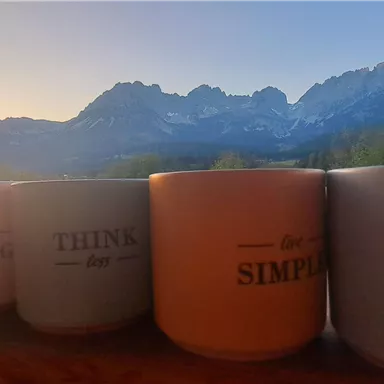 Four cups with positive words are on a table as the sun sets behind majestic mountains. The sky is colorful and conveys a peaceful atmosphere.