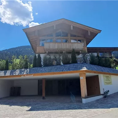 A modern house with wooden elements and a large balcony. In the background, green meadows and mountains can be seen.