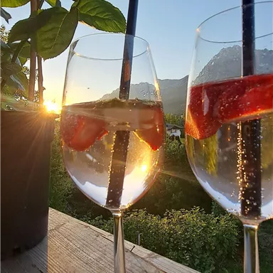 Two glasses with drinks are sitting on a wooden tray, filled with water and strawberries. In the background, a beautiful sunset can be seen.