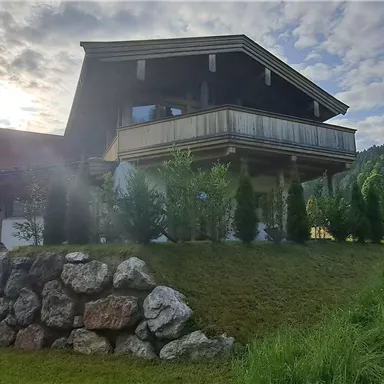A modern house on a hill, surrounded by greenery and mountains. The sun is shining behind the building, creating a beautiful atmosphere.