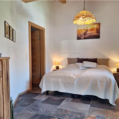 A cozy bedroom with a large bed and an elegant chandelier. On the wall hangs a picture of a stag, and the floor is made of stones.