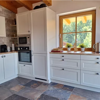 A modern kitchen with white cabinets and a large window. The countertop is neatly equipped with some kitchen utensils and decorative plants.