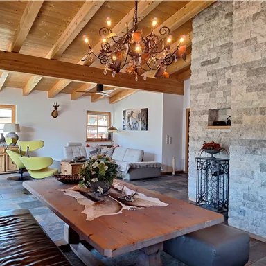 A cozy living room with wooden ceilings and a large dining table. The decor is modern with rustic elements and a beautiful stone wall.