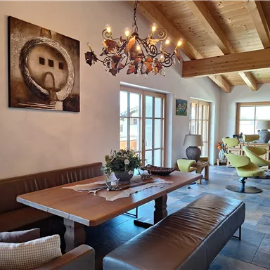 An inviting dining room with a large wooden table and cozy seating options. The room has bright walls, wooden ceilings, and a modern chandelier.