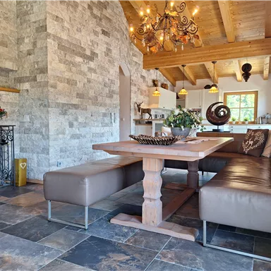 A modern dining room with a large wooden table and stylish benches. The walls are clad in stone, and a beautiful ceiling light hangs over the table.