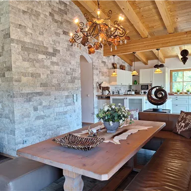 A cozy dining area with a large wooden table and seating. The walls are clad in natural stone, and there is a bright, inviting kitchen in the background.