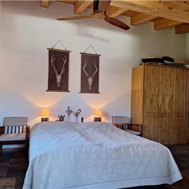 A cozy bedroom with a large bed and wooden furniture. Decorative pictures hang on the walls, and the lighting is warm and inviting.