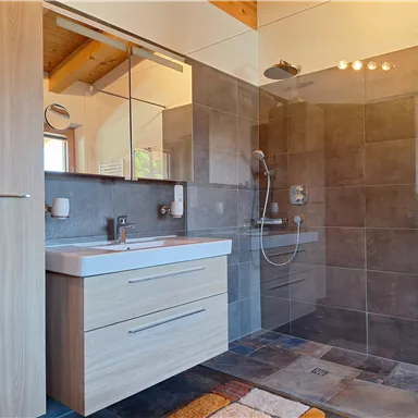 A modern bathroom with a spacious shower and wooden details. The sink area features a light wood dresser and large mirrors.