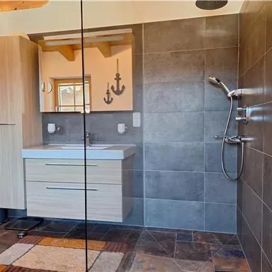 A modern bathroom with a shower and a sink in wood look. The walls are designed with gray tiles and there is a mirror.