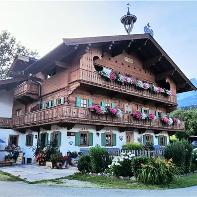 Ein traditionelles alpenländisches Haus mit Holzverkleidung und vielen Blumenbalkonen. Die Umgebung ist grün und sonnig, mit Bergen im Hintergrund.