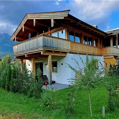 A modern house with wooden cladding and a balcony. The surroundings are green with trees and a beautiful mountain landscape in the background.