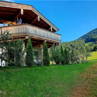 A cozy wooden house stands on a green meadow in the mountains. In the background, gentle hills stretch out under a blue sky.