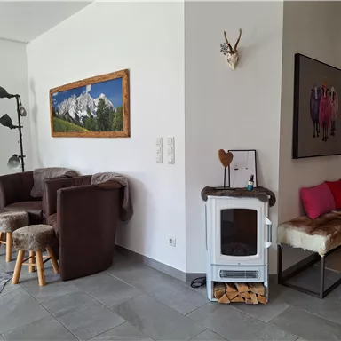 A cozy living space with modern furniture and a wood stove. On the wall hangs a picture of mountains, and there is a small seating area with cushions.
