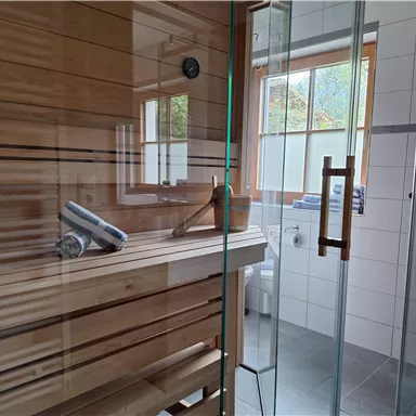 A modern sauna with wooden shelves and a large glass front. Brightly lit by windows, it offers a relaxing atmosphere.