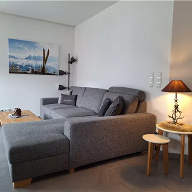 A cozy living room with a gray sofa and a small side table. On the wall hangs a picture of a mountain landscape with skis.