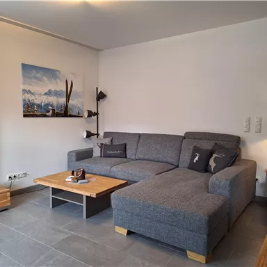 A modern living room with a large gray sofa and a wooden table. A landscape painting of the mountains hangs on the wall.