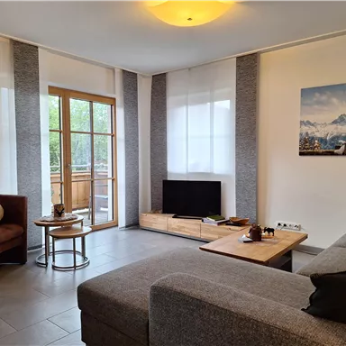 A modern living room with a gray couch and a small table. Large windows let in plenty of light and provide access to a balcony.