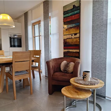 A modern dining area with a wooden table and chairs. A comfortable armchair and an artistic wall painting complement the cozy atmosphere.