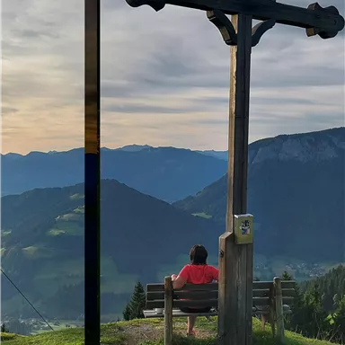 A person is sitting on a bench looking at a beautiful mountain landscape. In the background, there is a black cross and the sky is lightly cloudy.