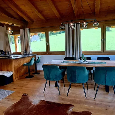 A cozy dining area in a wooden house with large windows and a view of the greenery. The room is modernly furnished with a dining table and stylish chairs.