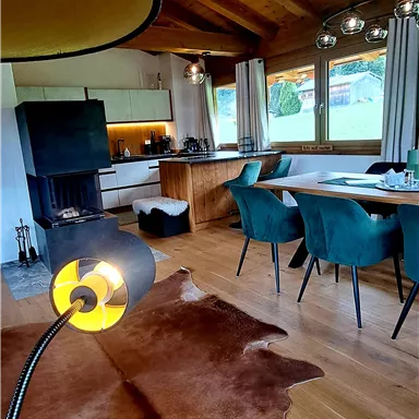 A modern living room with wooden beam ceiling and large windows. There is a stylish dining table and comfortable chairs, as well as a leather rug.