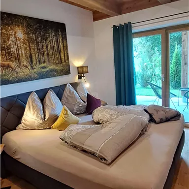 A cozy bedroom with a large bed and many pillows. Large windows offer a view of the green nature.