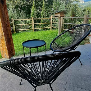 Two modern chairs and a small table are on a terrace. In the background, green trees and a fence can be seen.