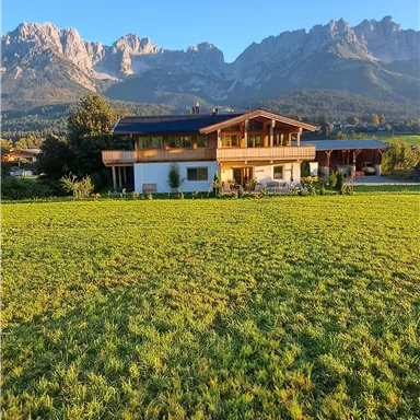A picturesque house on a meadow with the mountains in the background. The sky is clear and blue.