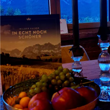 A beautiful table with a fruit bowl containing apples and grapes. In the background, there is a candle holder and a landscape photo of a mountain panorama.