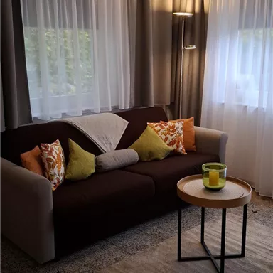 Ein gemütliches Wohnzimmer mit einem Sofa und bunten Kissen. Die Fenster sind mit hellen Vorhängen bedeckt, und ein kleiner Tisch steht davor.