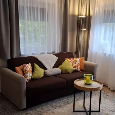 Ein gemütliches Wohnzimmer mit einem beigen Sofa und bunten Kissen. Es gibt einen kleinen Tisch mit einer Tasse und große Fenster mit Vorhängen.