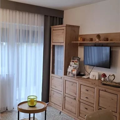 Ein gemütlicher Wohnraum mit Holzmöbeln und einem Fernseher. Helles Licht strömt durch die Vorhänge und sorgt für eine einladende Atmosphäre.