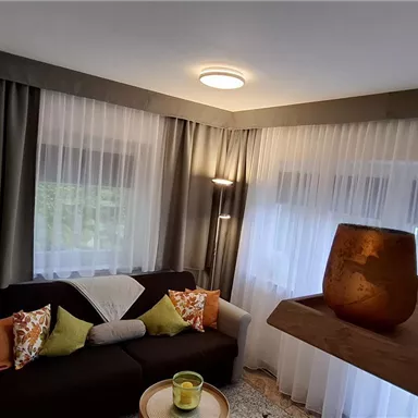 Ein gemütliches Wohnzimmer mit einem Sofa und bunten Kissen. Große Fenster lassen viel Licht herein und schaffen eine einladende Atmosphäre.