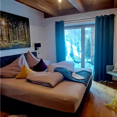 A cozy bedroom with a large bed and a wooden beam ceiling. Large windows offer a view of the outside and enhance the bright, inviting atmosphere.