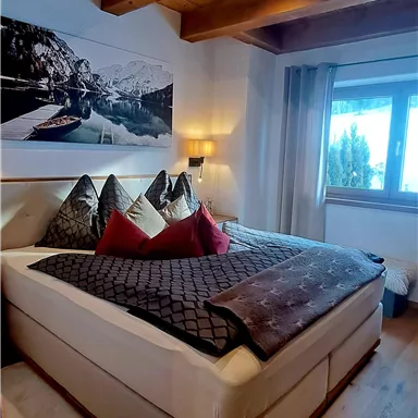 A cozy bedroom with a large bed and striking pillows. The wooden beam ceiling and the large window create an inviting atmosphere.