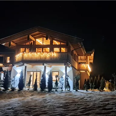 A beautiful, illuminated chalet at night, surrounded by snow. The trees in the foreground create a cozy atmosphere.