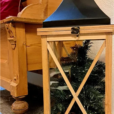 A wooden lantern stands next to a wooden bench and contains a small artificial fir tree. The room has a cozy atmosphere with warm colors.